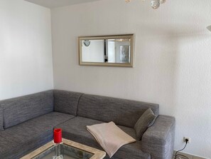 Ein Wohnzimmer mit einem grauen Sofa, einem kleinen Glastisch, einer roten Kerze und einem beigen Zierkissen.
