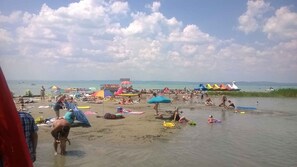 A beach with people sunbathing, swimming, and playing in the water. There are colorful umbrellas and a boat in the background. - Holiday apartment only 250 m from the sandy beach of the Golden Shore (Siófok)
