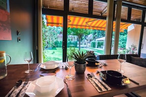 A dining table set with plates, cutlery, and wine glasses, with a view of a garden and a house through large windows. - Holiday apartment only 250 m from the sandy beach of the Golden Shore (Siófok)