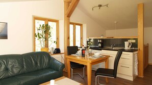 A cozy living room with a green leather sofa, a wooden dining table with chairs, a kitchen area with a stove, and a large window with a plant. - Pfarrweg 10 PF-10 - PF-10 (Kühlungsborn)