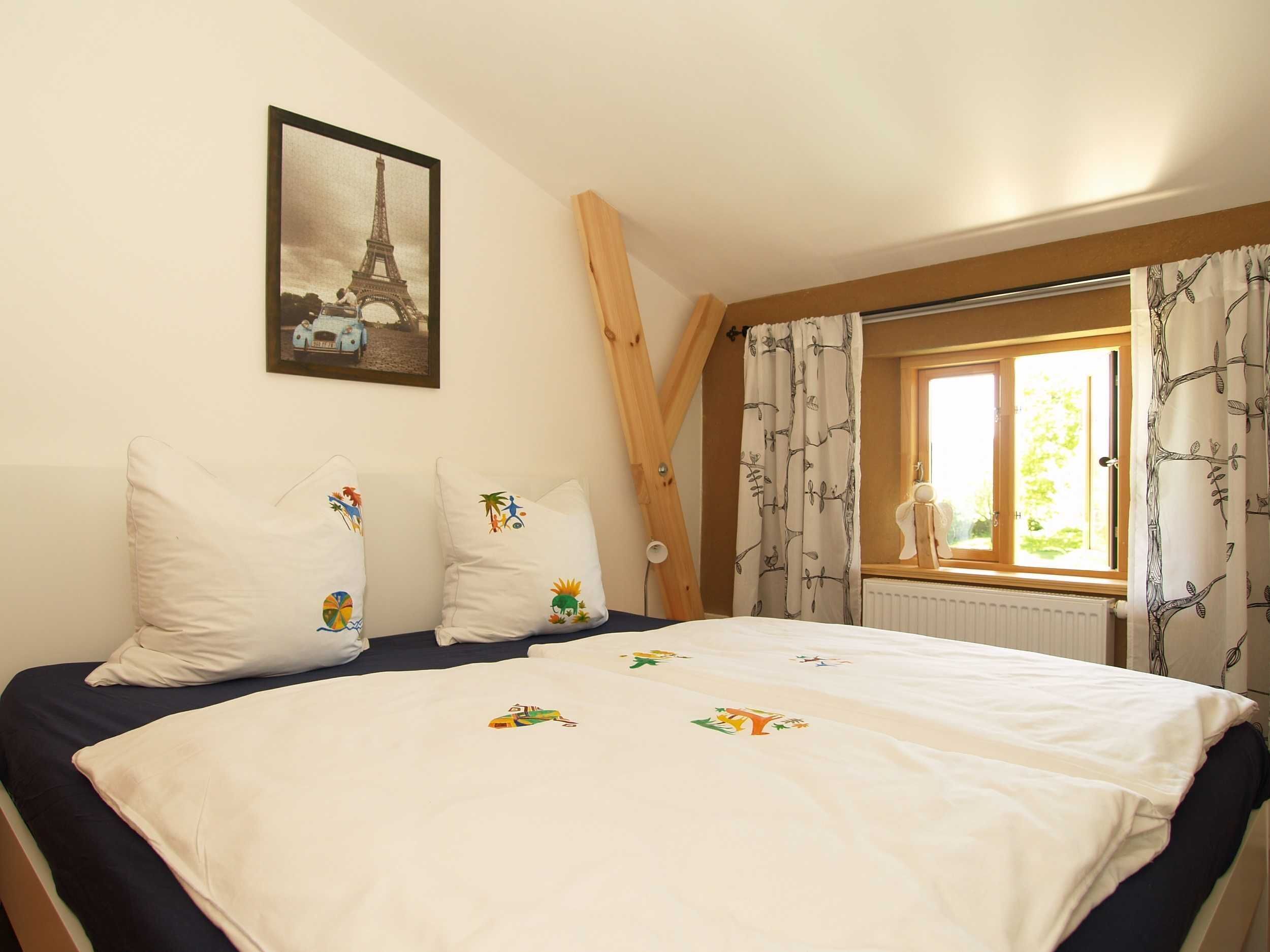 Ein Schlafzimmer mit einem Bett, Kissen mit bunten Mustern, einem Fenster mit Vorhängen und einem gerahmten Bildstücken vom Eiffelturm an der Wand.