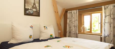 Ein Schlafzimmer mit einem Bett, Kissen mit bunten Mustern, einem Fenster mit Vorhängen und einem gerahmten Bildstücken vom Eiffelturm an der Wand.