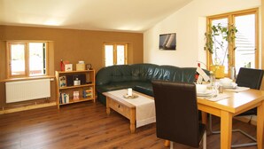 Ein gemütliches Wohnzimmer mit einem grünen Ledersofa, einem Holztisch und einem Esstisch mit Stühlen. Zwei Fenster sorgen für natürliches Licht, ein Heizkörper ist vorhanden, und ein Bücherregal zeigt verschiedene Gegenstände.