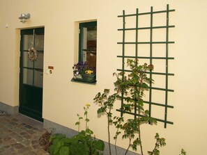 Una entrada de casa con una puerta verde, un ventanal pequeño con macetas de flores y una celosía verde en la pared.