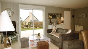Una sala de estar con sofá, reposapiés, mesa de centro y televisor. Se observa una ventana con vista a una casa y árboles.