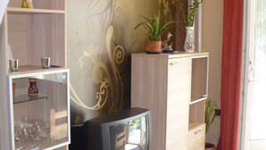 Uma sala de estar com televisão sobre um suporte de madeira, parede decorativa com padrão floral e uma planta em vaso numa prateleira.