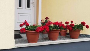 Entrada de um edifício com porta branca e parede amarela. Cinco vasos de terracota com flores vermelhas estão nos degraus.