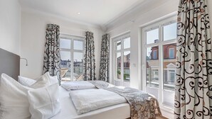 A hotel room with a large bed, white bedding, patterned curtains, and a view of buildings through large windows. - Villa Aquamarina Whg.26a, ROOF TERRACE, LAKE VIEW, CHAIR - Villa Aquamarina, 1st floor. (Heringsdorf)