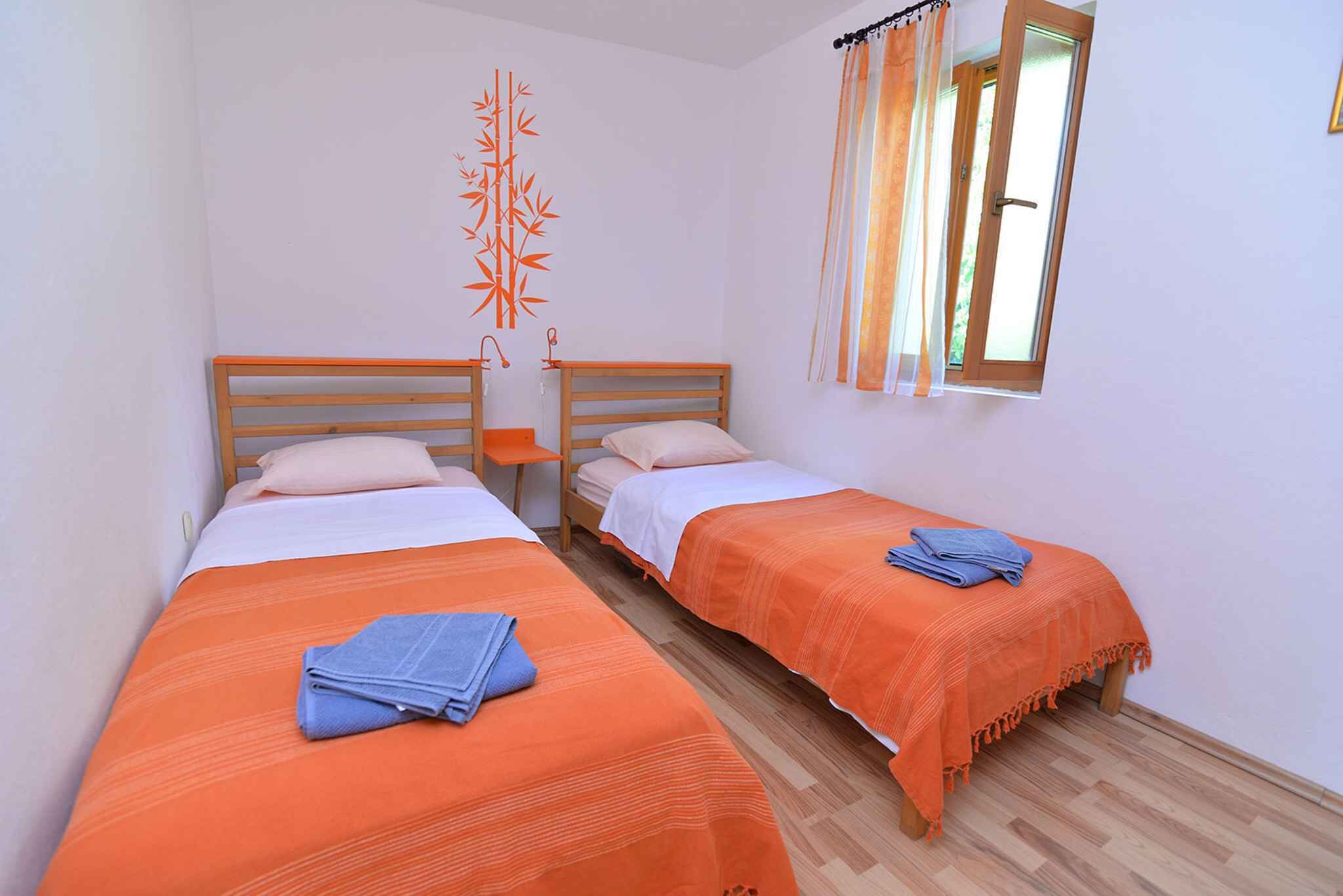 Ein Zimmer mit zwei Betten, jeweils mit einer orangefarbenen Tagesdecke und einem blauen Handtuch. Ein Fenster mit Vorhängen und eine Wanddekoration über den Betten sind vorhanden.