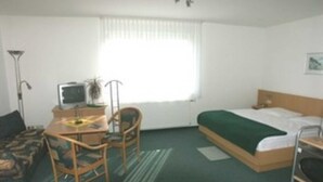 A hotel room with a bed, a desk with a computer, a chair, and a window with curtains. - App. 28 - Apartment house Anne (Binz)