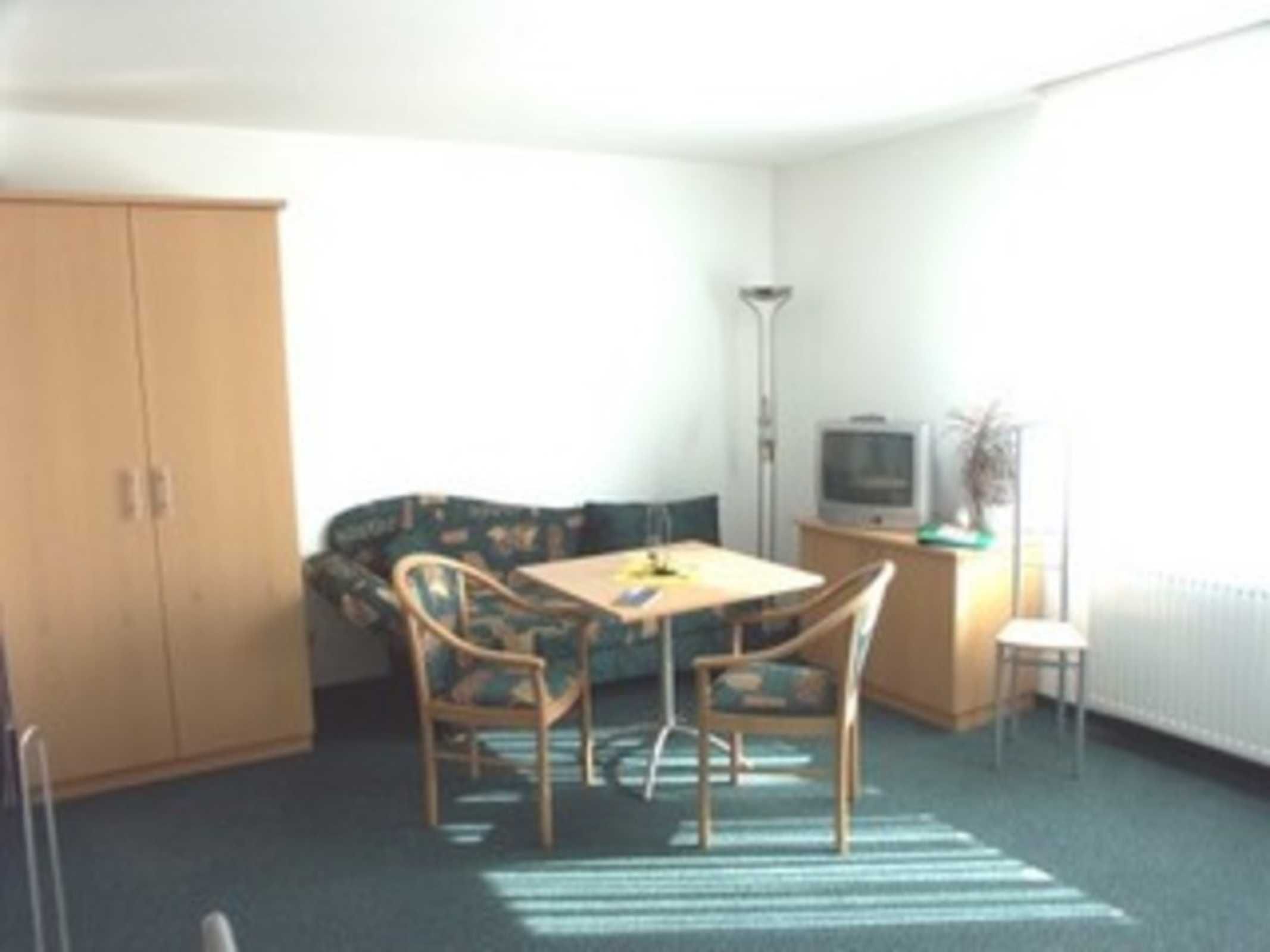 A small, sparsely furnished room with a single table and two chairs, a sofa, a cabinet, and a television.