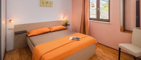 Une chambre à coucher comprenant un lit, une table de chevet, une chaise, une fenêtre avec des rideaux et une étagère murale agrémentée d’une plante.