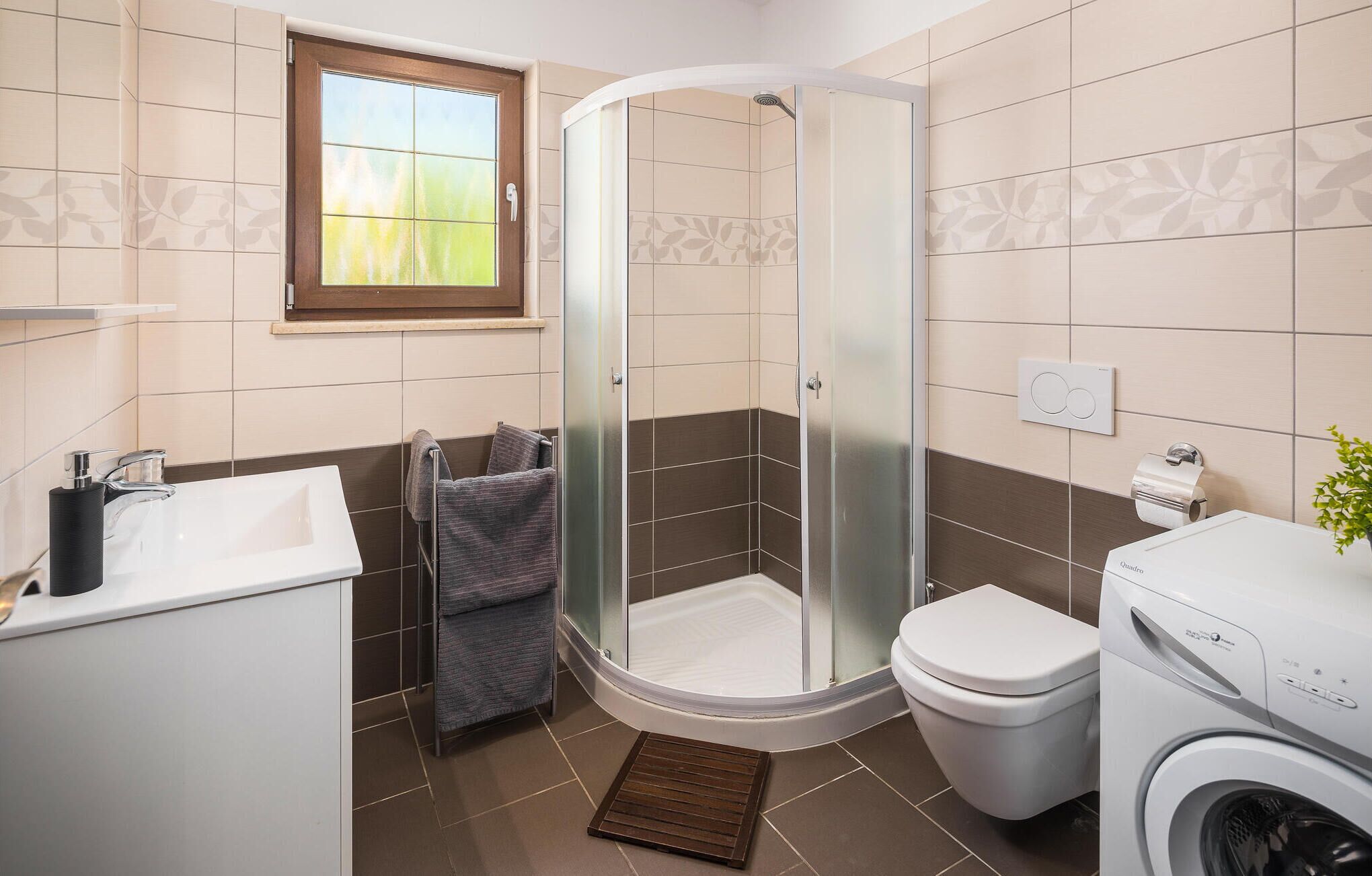Une salle de bain moderne avec une douche à parois de verre, des toilettes, un lave-linge et une fenêtre donnant sur un espace verdoyant.
