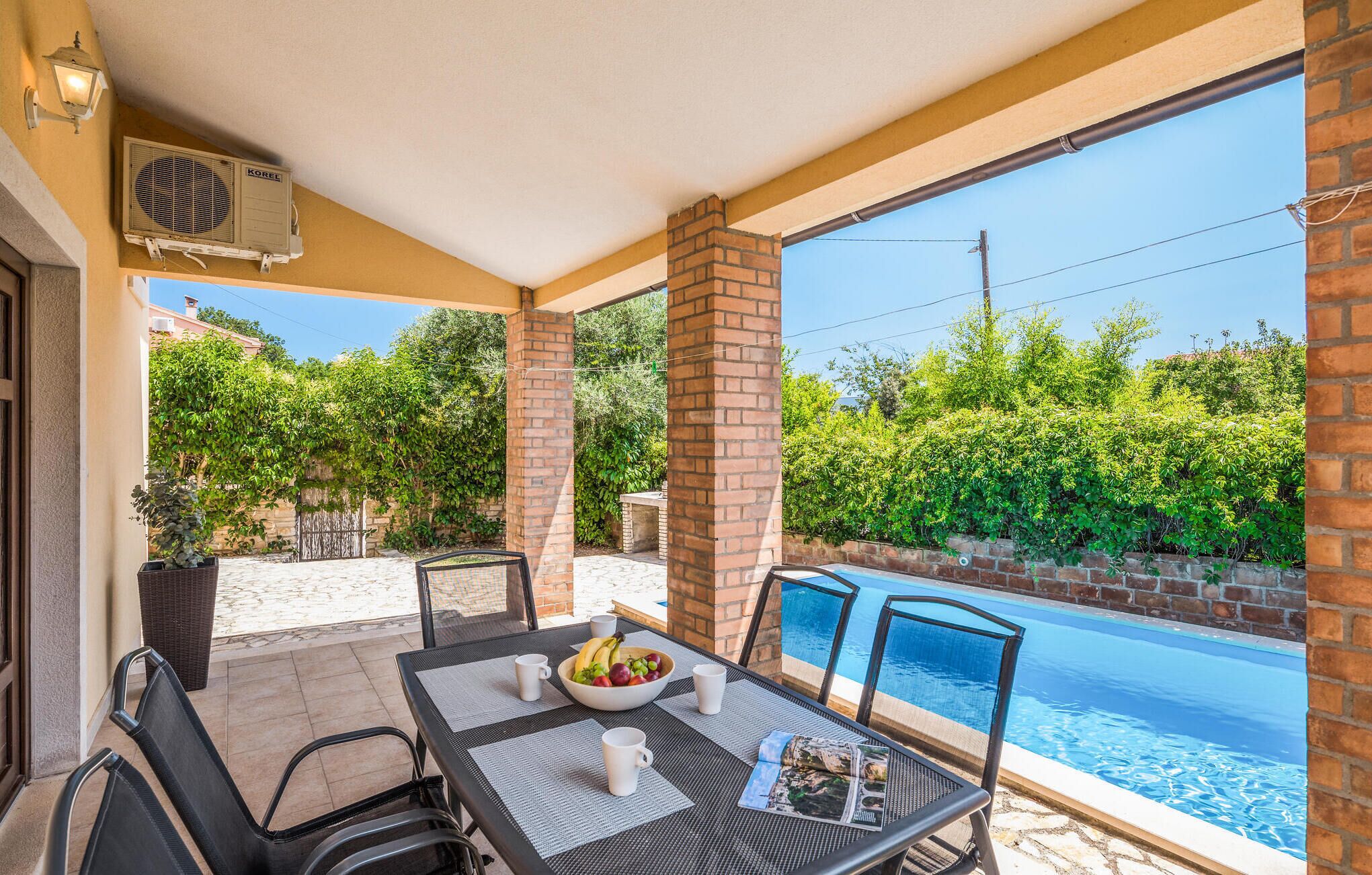 Un espacio exterior techado con vista a la piscina, que incluye una mesa con sillas, un cuenco de fruta y una revista.