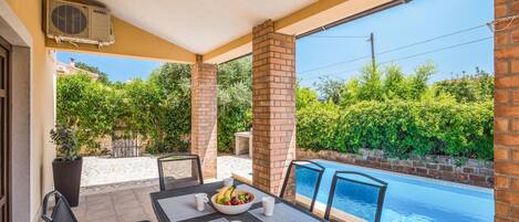 Un espacio exterior techado con vista a la piscina, que incluye una mesa con sillas, un cuenco de fruta y una revista.