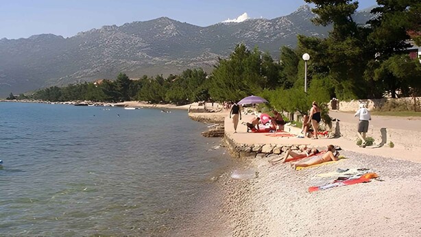 Une plage à l’eau claire, bordée de galets, avec des montagnes en arrière-plan.