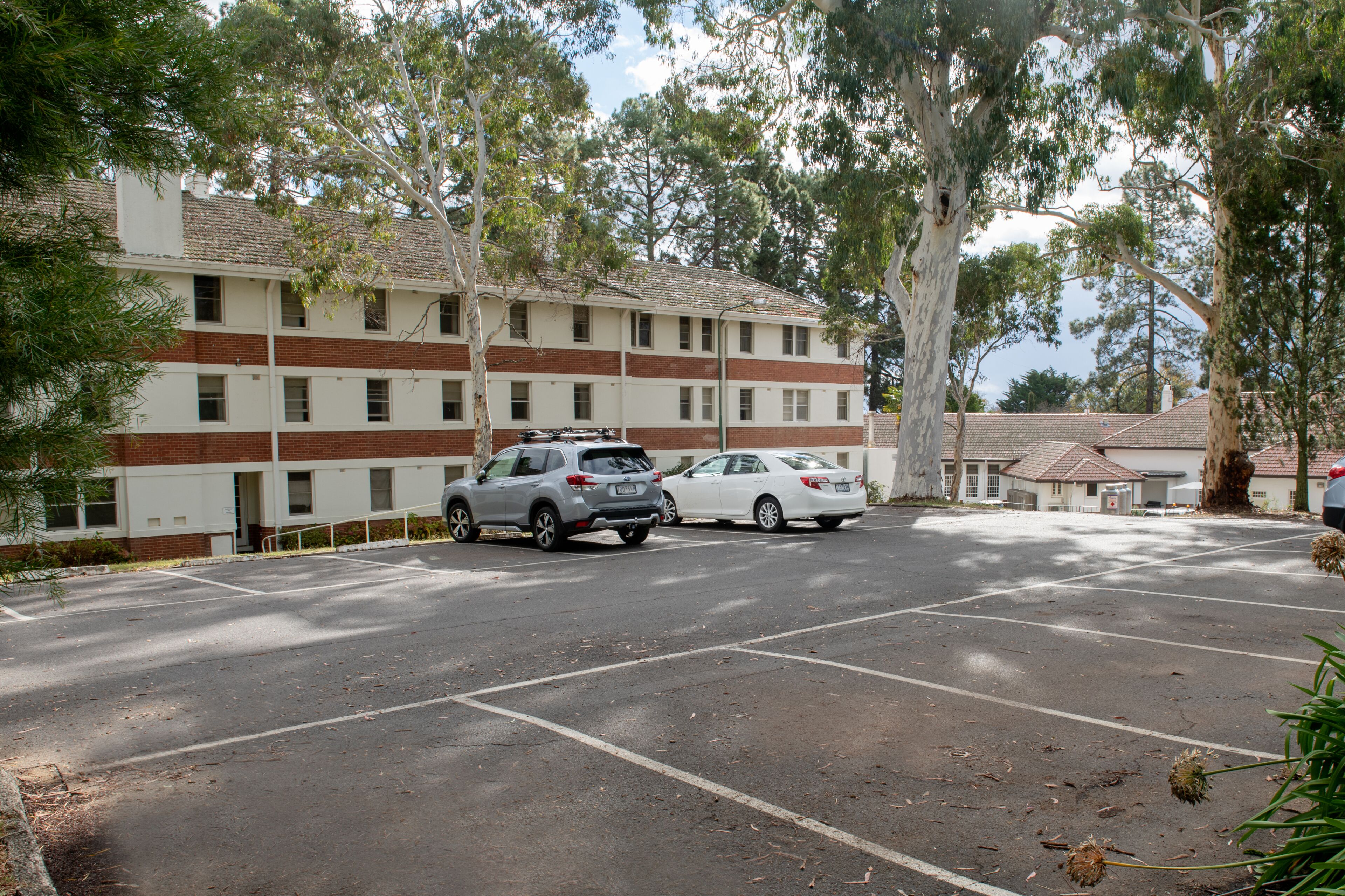 A parking lot with two cars, a building with multiple windows and a brick facade, and surrounding trees.