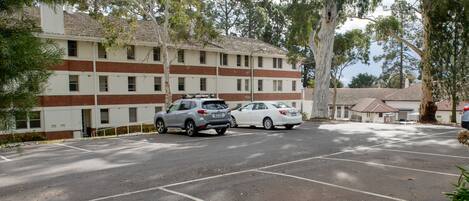 A parking lot with two cars, a building with multiple windows and a brick facade, and surrounding trees.