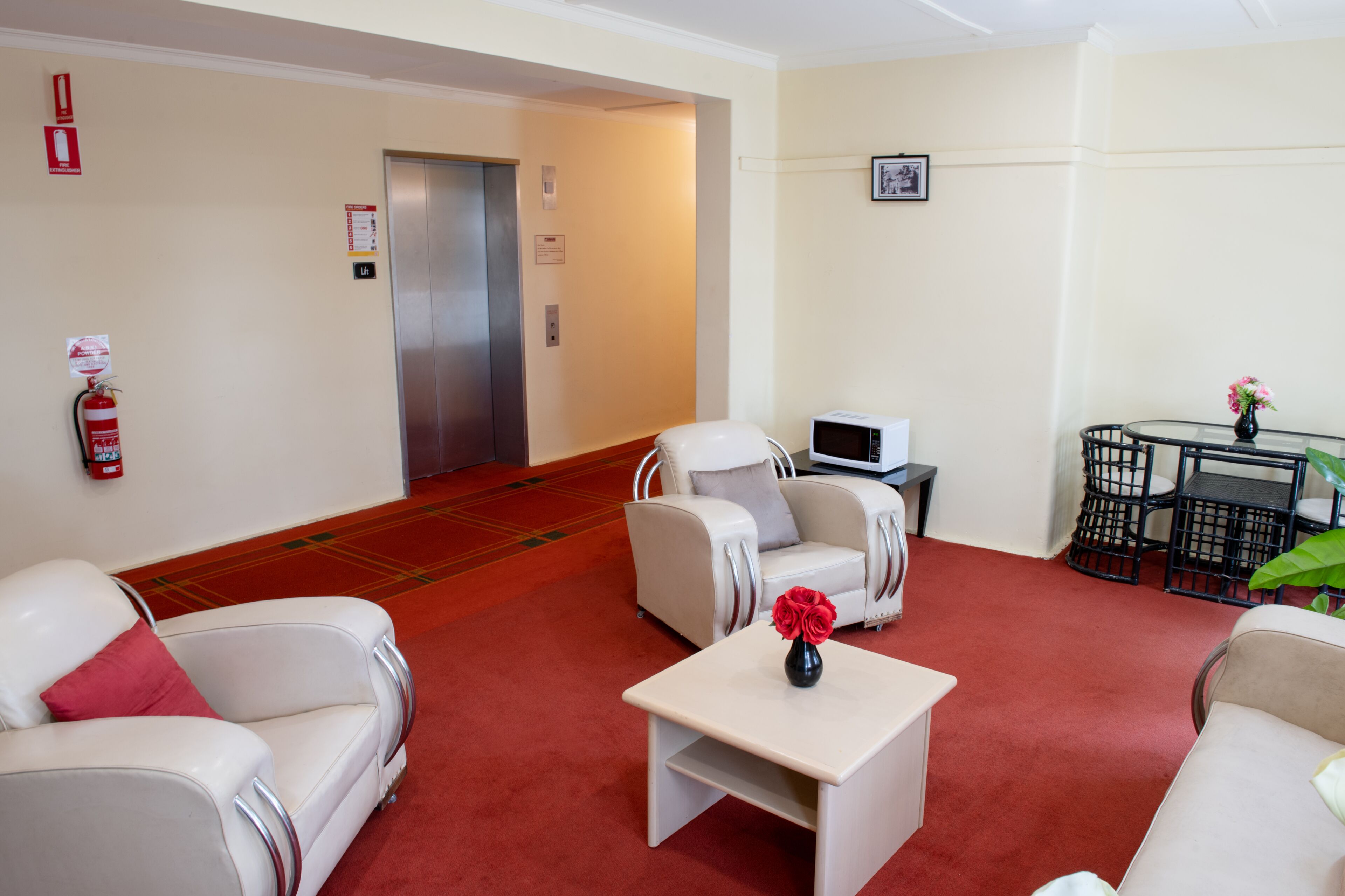 A hotel lobby with a red carpet, white sofas, a small table with a vase, a microwave, and a fire extinguisher.