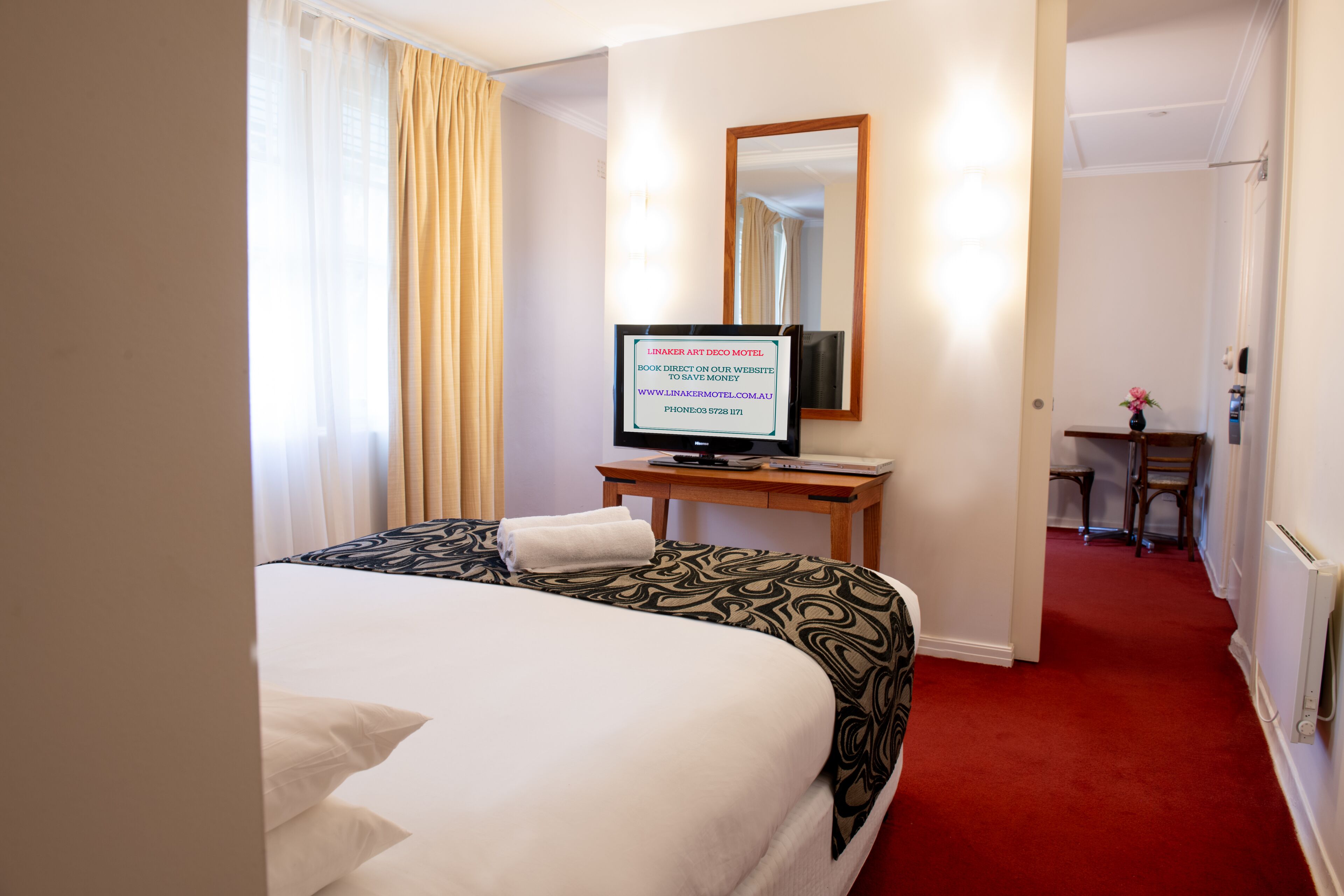Habitación de hotel con cama, televisión sobre una mesa de madera, espejo en la pared y pasillo con alfombra roja.