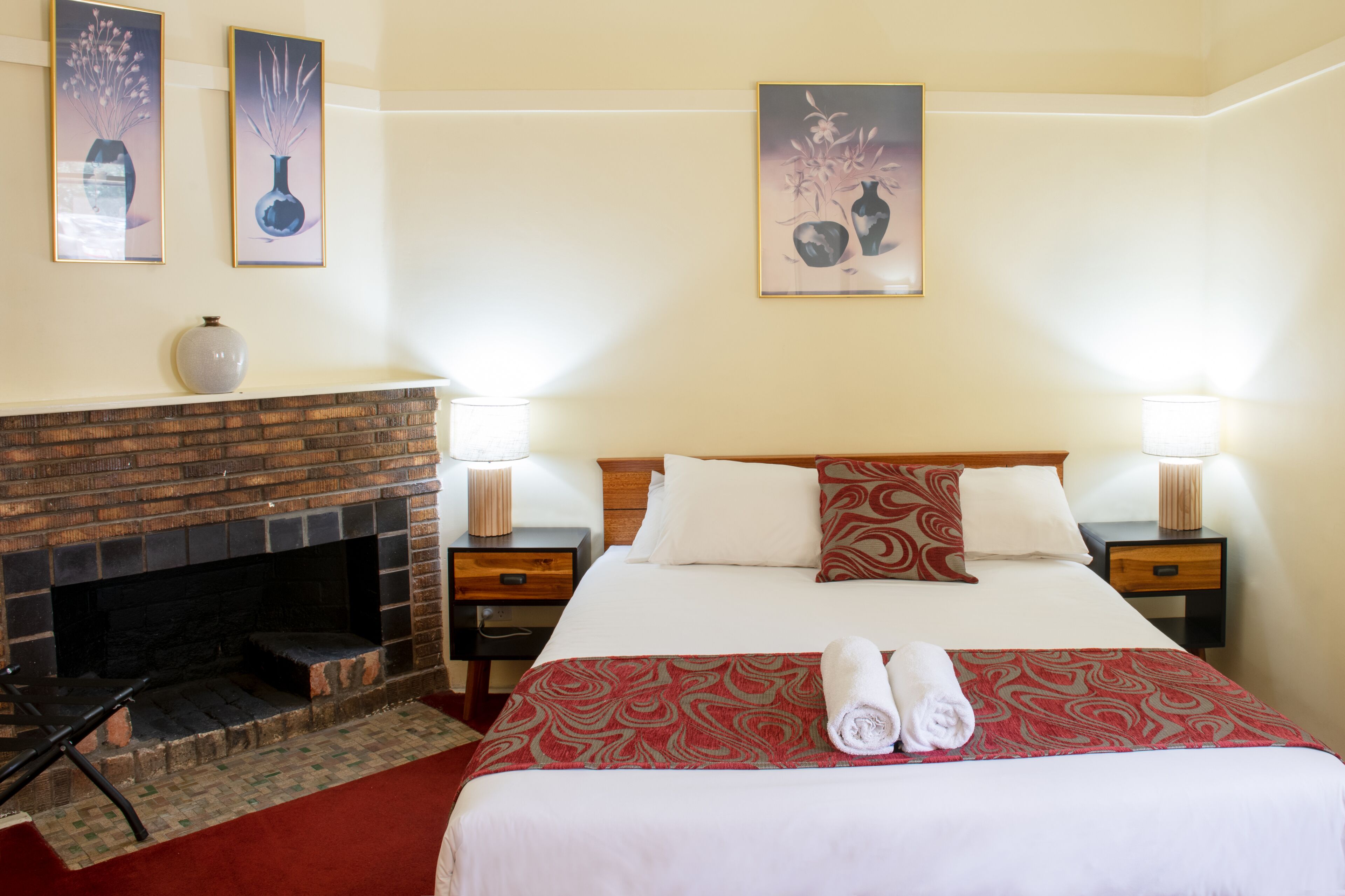 Habitación de hotel con chimenea de ladrillo, una cama grande con ropa de cama roja y blanca, dos mesitas de noche con lámparas, y tres obras de arte enmarcadas en la pared.