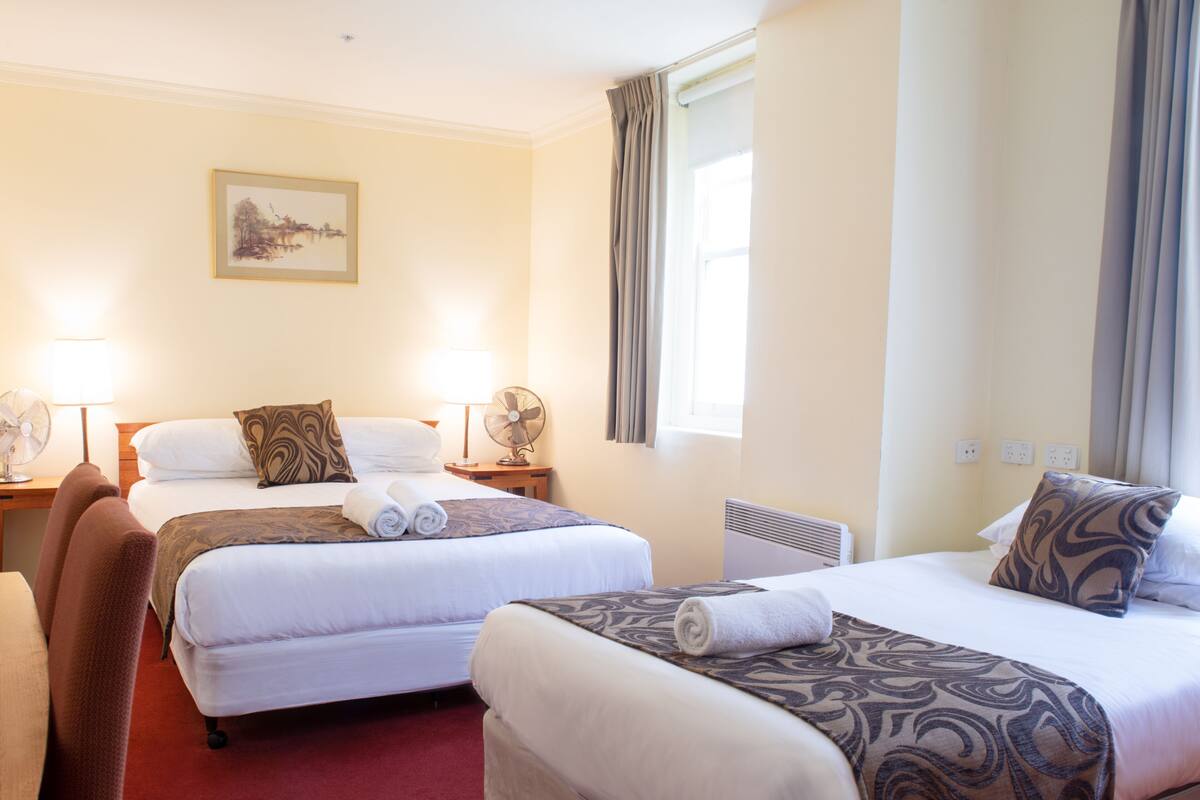 A hotel room with two beds, a sitting area, a fan, a lamp, and a painting on the wall.