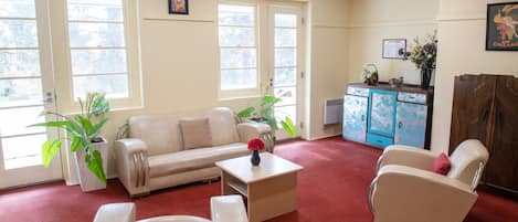 A spacious living room with a beige sofa, a white armchair, and a coffee table. There are large windows allowing natural light, and a blue cabinet in the background.
