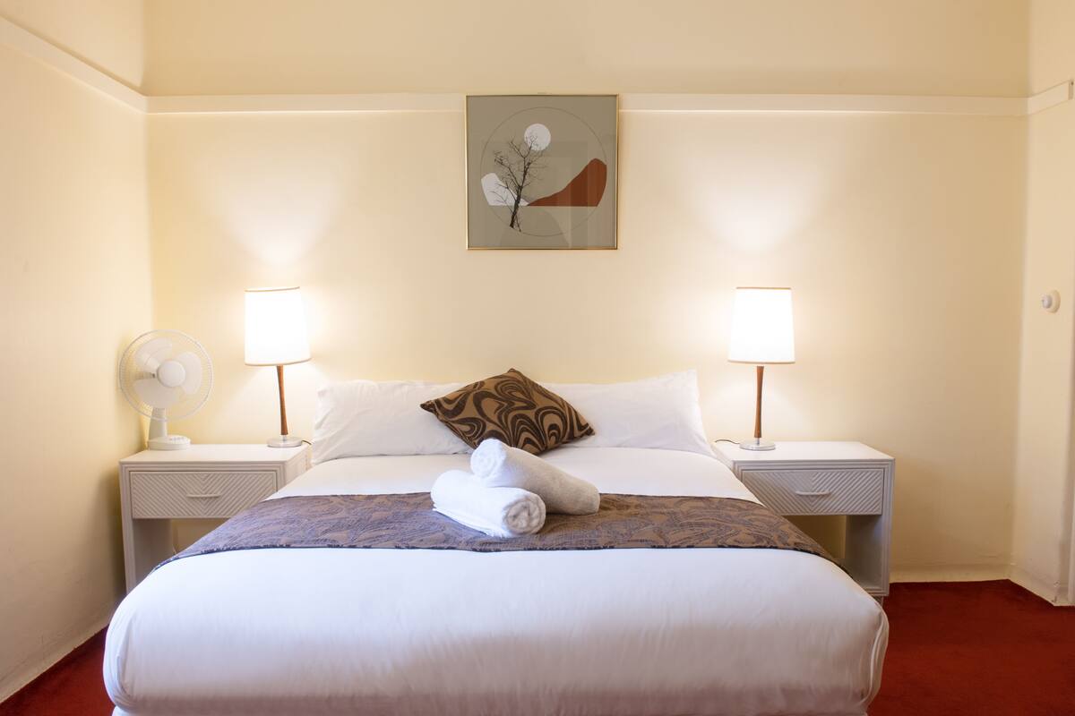 A neatly made bed with white linens, two bedside lamps, a wall-mounted artwork, and a white nightstand with a fan.