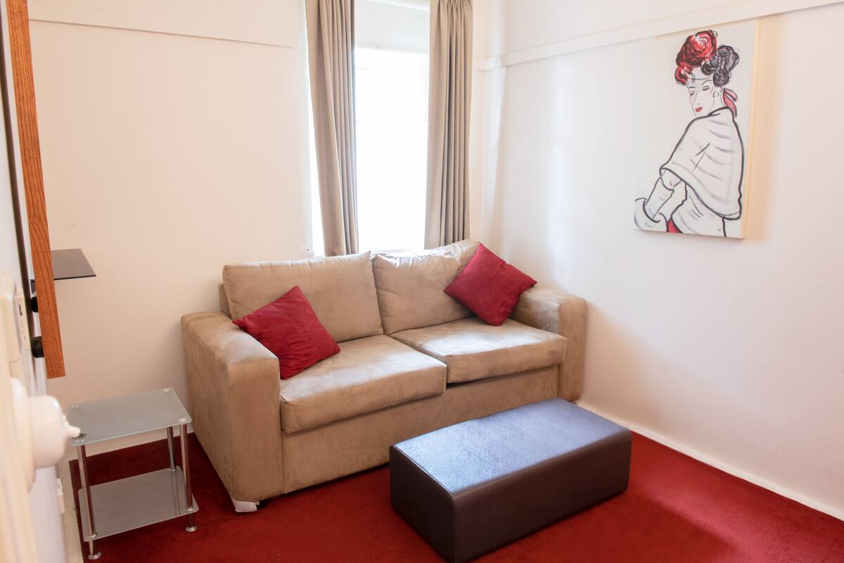 A living room with a beige sofa, red cushions, a small side table, a black ottoman, and a wall-mounted artwork.