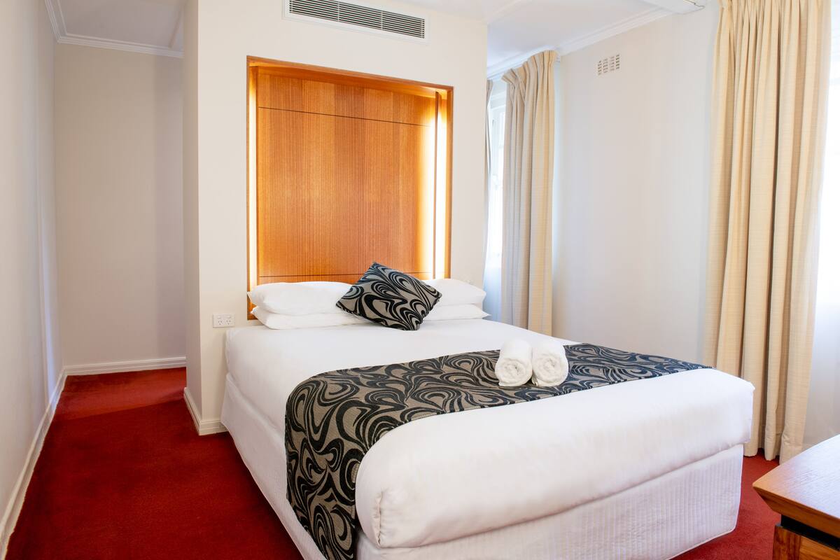 A hotel room with a bed, a wooden headboard, a window with curtains, and a red carpet.