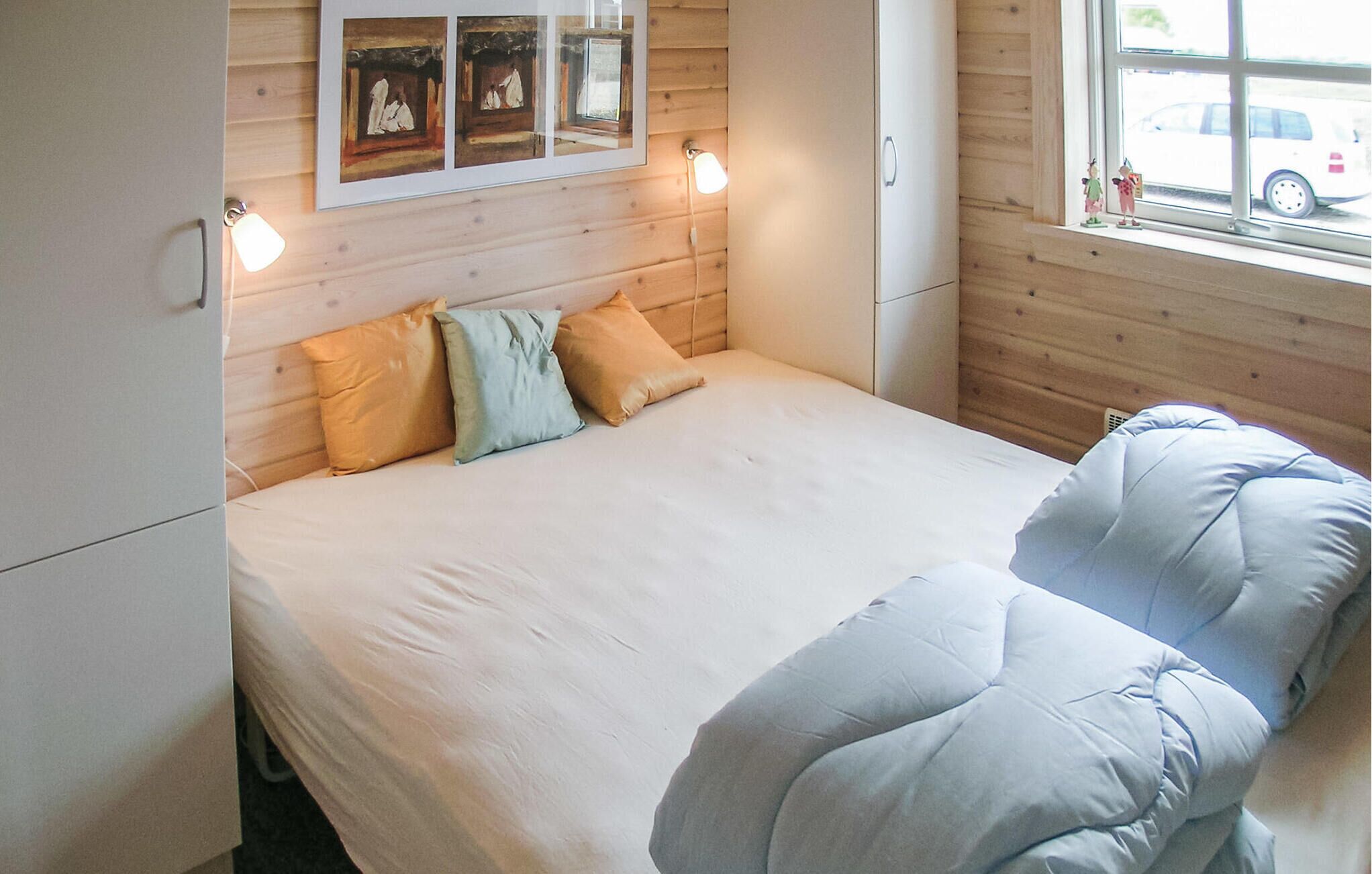 A bedroom with a bed, pillows, a headboard, a window with a view of parked cars, and a wall-mounted picture.