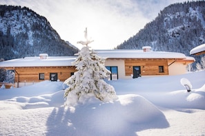 A wooden cabin with a snow-covered roof, surrounded by snow-covered trees and mountains.