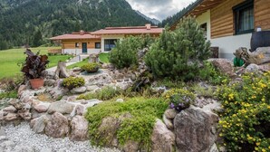 A wooden cabin with a red roof, surrounded by a well-maintained garden with various plants and a stone pathway leading to the entrance. - Chalet Holiday House 12 - Almdorf Tirol (Haldensee)