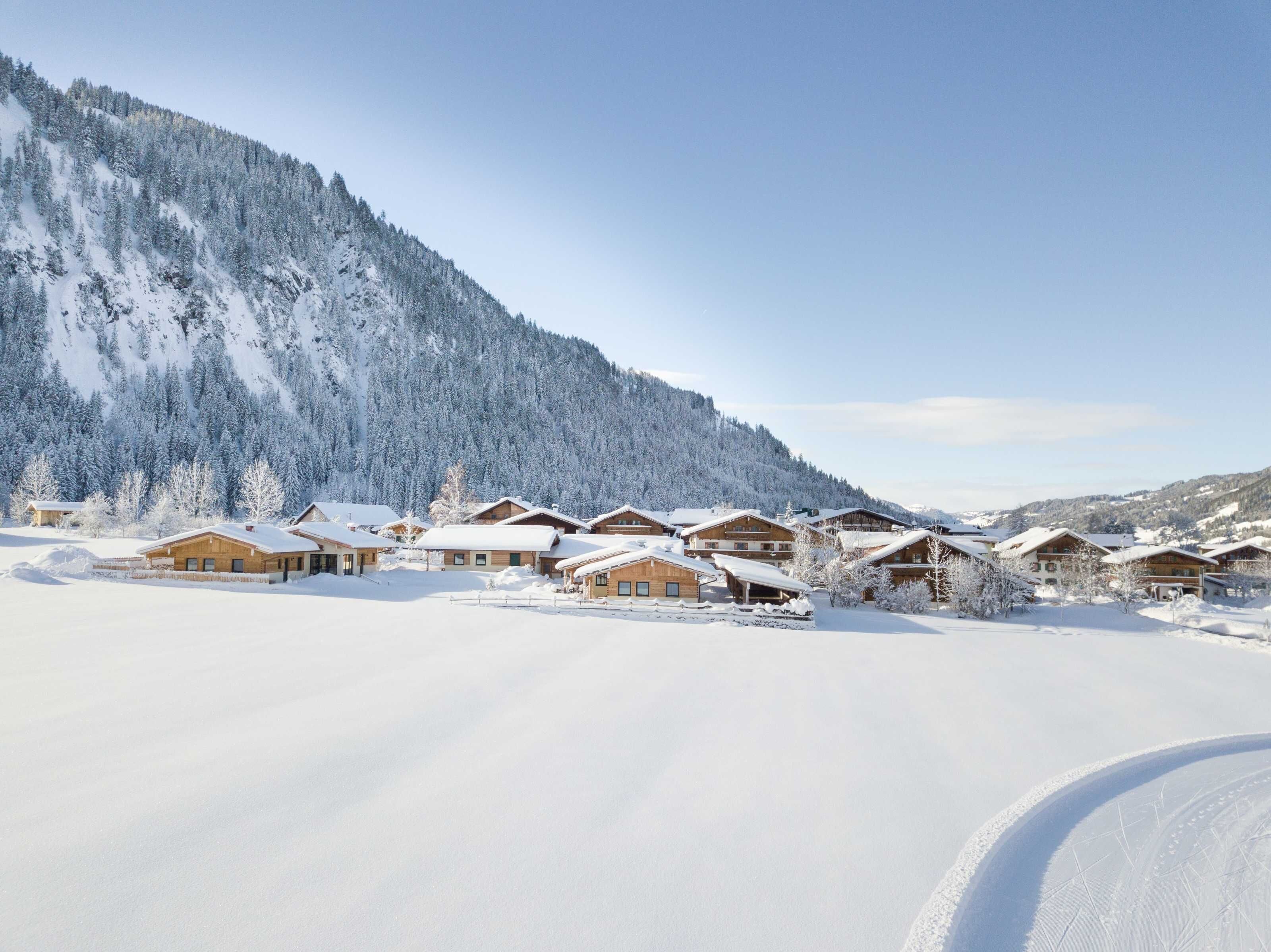 A snow-covered mountain village with wooden chalets, a clear sky, and a ski slope.