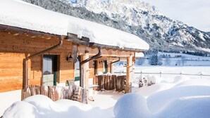 A wooden cabin with a snow-covered roof, a wooden porch, and a view of snow-covered mountains in the background. - Chalet Holiday House 12 - Almdorf Tirol (Haldensee)