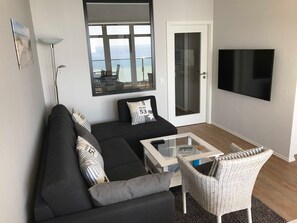 Ein modernes Wohnzimmer mit einem schwarzen Sofa, einem Glastisch, einem Korbstuhl, einem Flachbildfernseher und einem großen Fenster mit Meerblick.