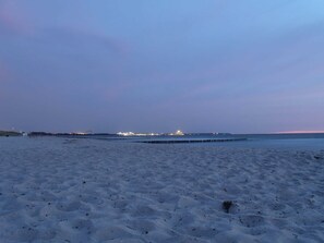 A beach with a pier extending into the water, a calm sea, and a city skyline in the distance.