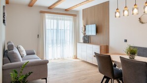 A modern living room with a sofa, dining table, and chairs, wooden beams on the ceiling, and a TV on the wall. - Sunnhof - Arnika vacation apartment (Gegend)