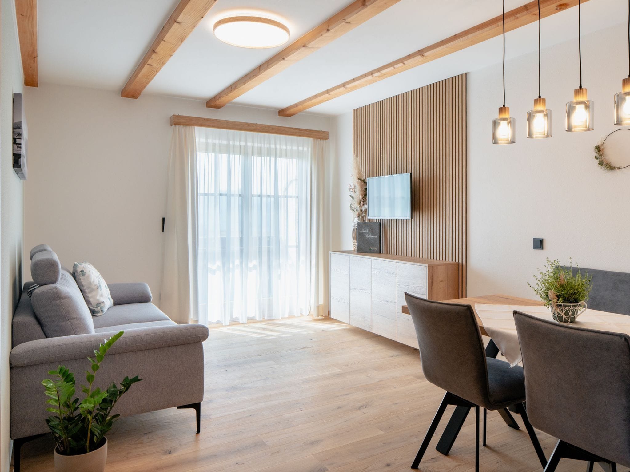 Ein modernes Wohnzimmer mit einer Couch, einem Esstisch, Stühlen, Holzbalken an der Decke und einem Fernseher an der Wand.