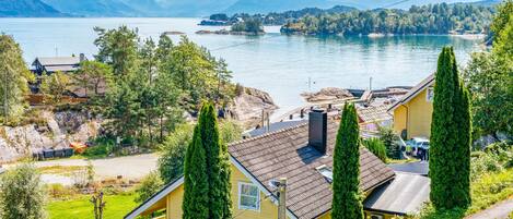 A yellow house with a sloped roof, surrounded by greenery and a view of a lake and mountains.