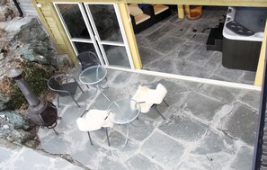 Een terras met stenen vloer, metalen stoelen, een klein tafeltje en een zithoek met kussens.