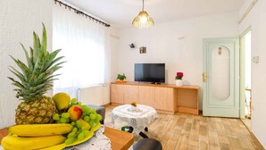 Un petit salon lumineux avec un plancher en bois, un téléviseur posé sur un meuble en bois, une table à manger ornée de fruits et une porte de couleur vert clair.
