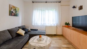 Un salon avec un canapé gris, une petite table ronde recouverte d’une nappe en dentelle, un téléviseur posé sur un meuble en bois et une fenêtre ornée de rideaux légers.