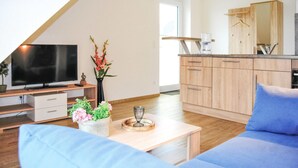 Una sala de estar moderna con televisor, sofá y mesa de centro. Hay una cocina con gabinetes de madera y una estante colgante.
