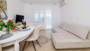 A modern living room with a white sofa, a dining table with chairs, a TV, and a large window. - Holiday apartment with terrace and sea view (Crikvenica)