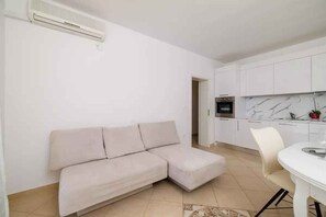 Een woonkamer met een beige bank, een eettafel met stoelen, een keuken met witte kasten en een marmeren achterwand, en een wit plafond.