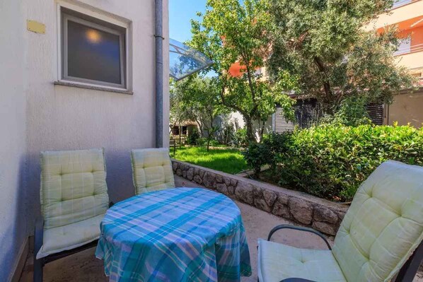 A patio with two chairs and a table, a window, and a garden with trees and bushes.