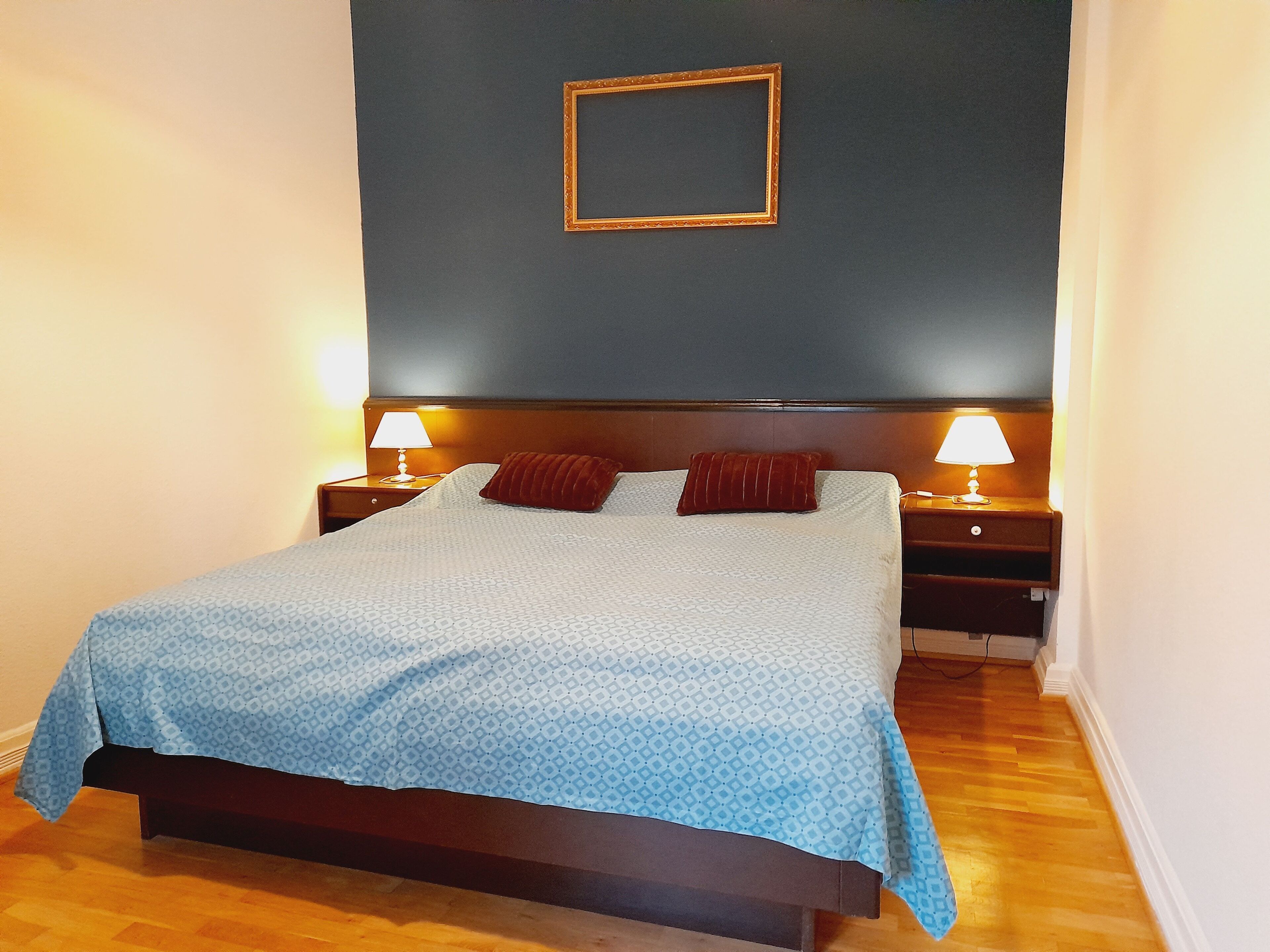 Un lit bien fait, recouvert d’une couvre-lit bleue à motifs, avec deux oreillers rouges et deux lampes de chevet posées sur des tables de nuit en bois.