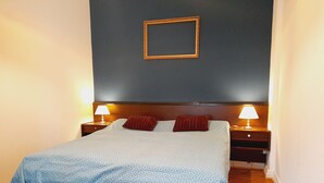 Un lit bien fait, recouvert d’une couvre-lit bleue à motifs, avec deux oreillers rouges et deux lampes de chevet posées sur des tables de nuit en bois.