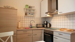Une cuisine moderne équipée d’un lave-vaisselle, d’un four et d’une machine à café. On y trouve une table blanche avec une chaise, ainsi qu’une étagère contenant des ustensiles de cuisine.